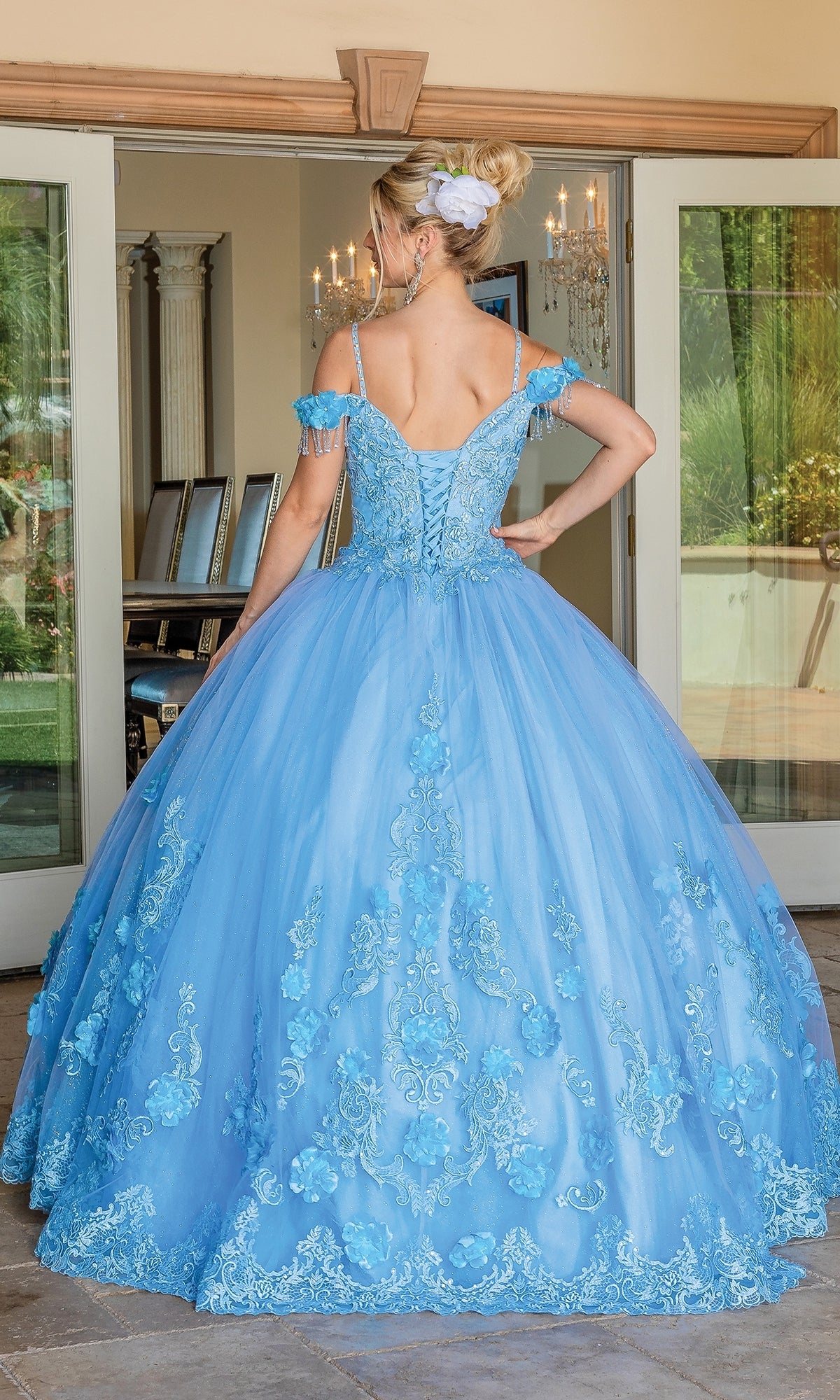 A woman faces away from the camera indoors, wearing the Quinceanera Dress 1706 by Dancing Queen—an ornate, light blue ball gown with floral embroidery and an off-the-shoulder design, standing in front of glass doors.